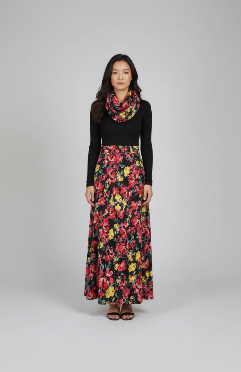 Woman wearing a black top, colorful floral skirt, and scarf on a gray background