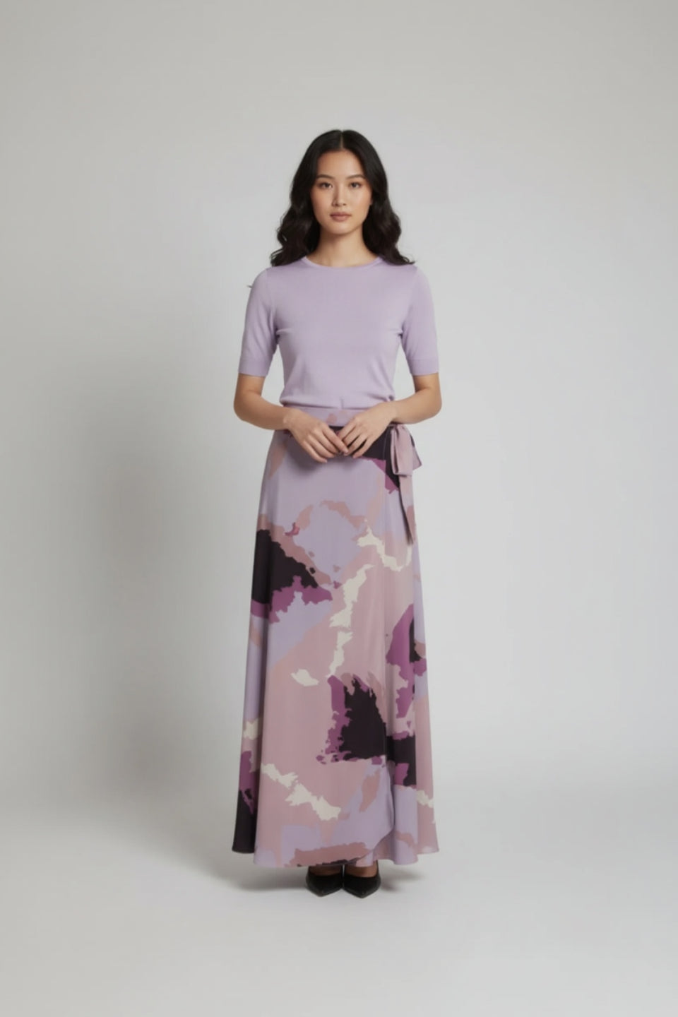 Woman wearing a light purple top and a long, patterned skirt on a plain background