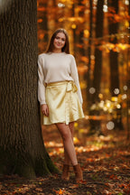 Woman in a yellow skirt and white sweater standing next to a tree in an autumn forest.
