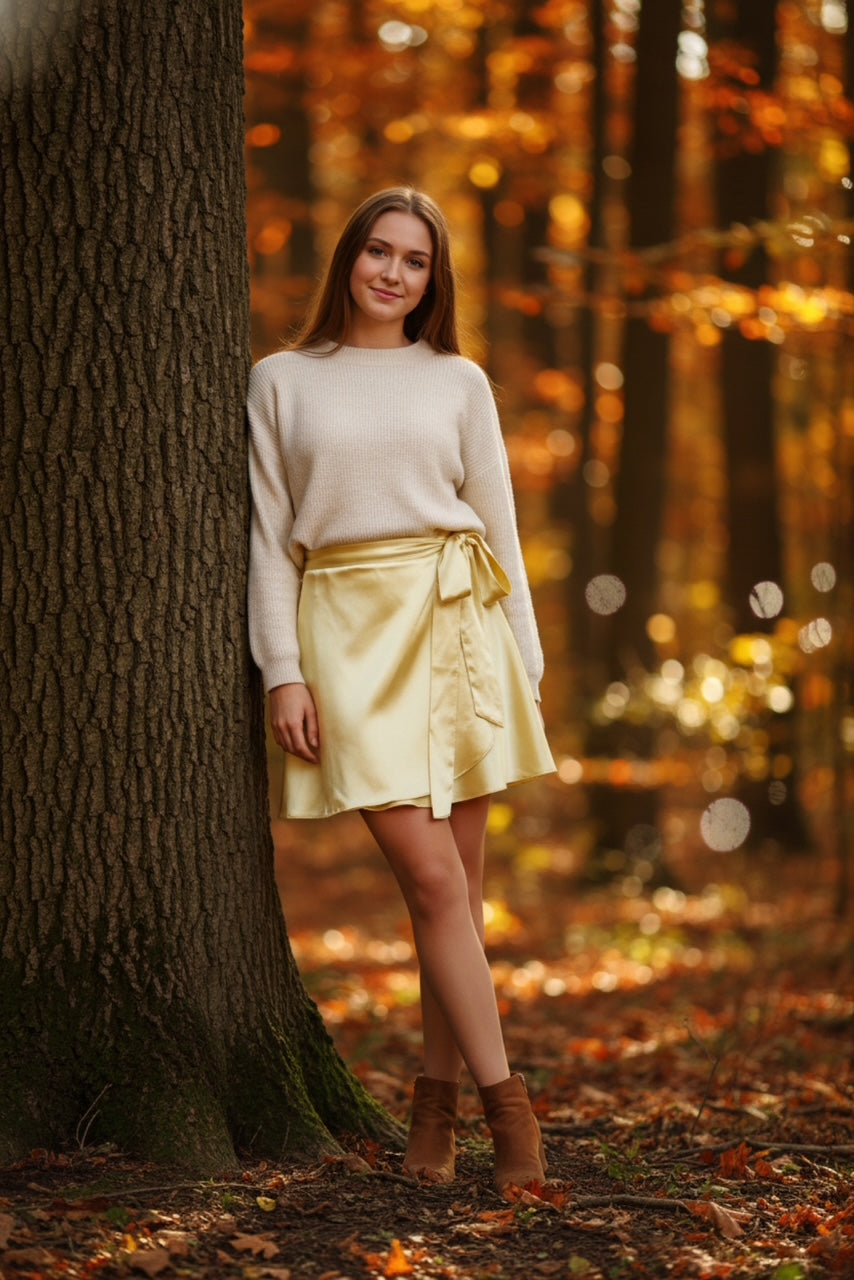 Woman in a yellow skirt and white sweater standing next to a tree in an autumn forest.